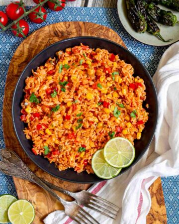 Orange rice with corn, tomato, and parsley in a black bowl. Lime slices are on the side.