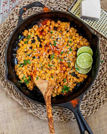 Cast iron skillet with charred yellow corn covered in spices and chopped herbs. A wooden spatula and two lime slices are placed inside.