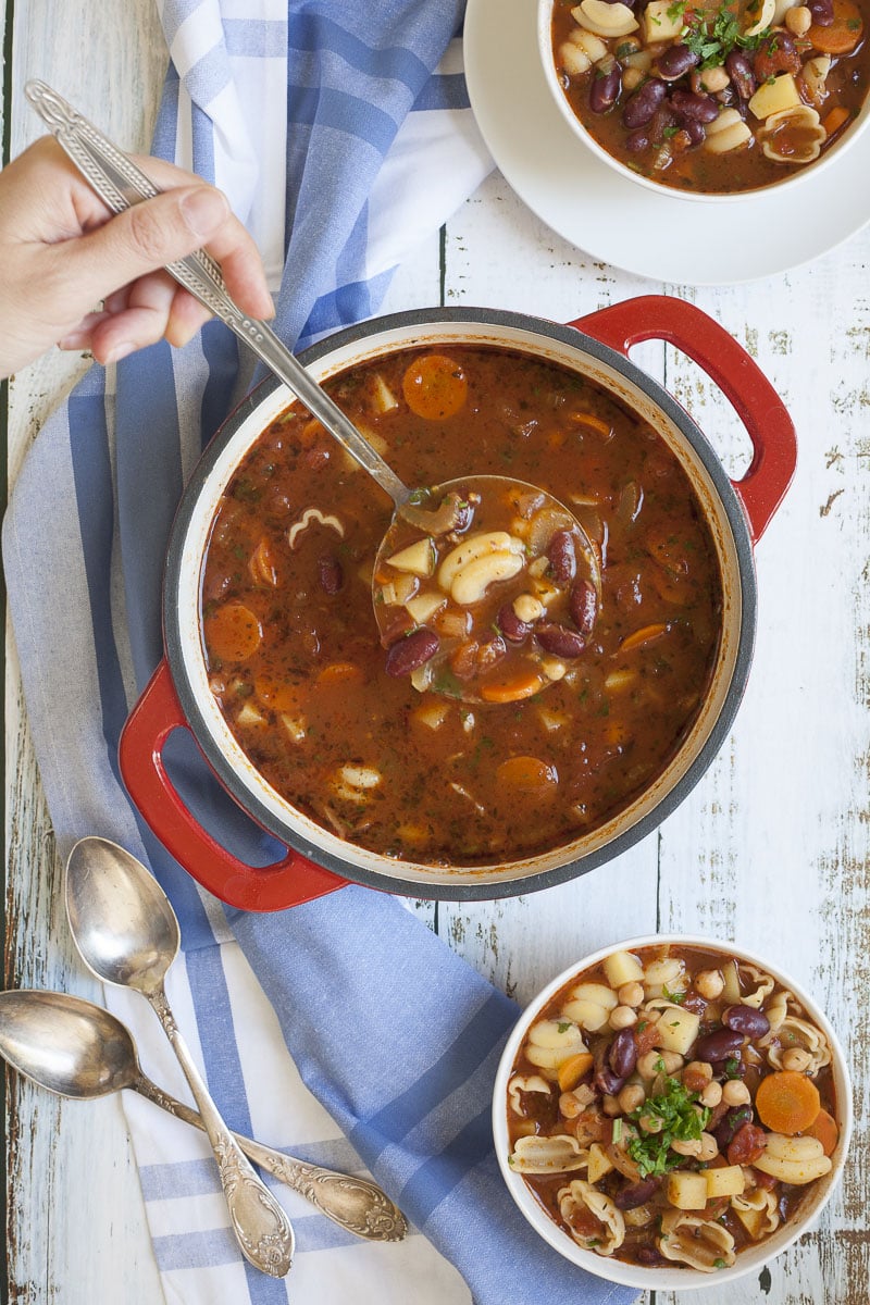 Red soup in a white dutch oven full of carrot slices, diced potatoes, beans, chickpeas and chopped green herbs. A hand holding a ladle is taking a serving. 2 white bowls are already full next to ti.