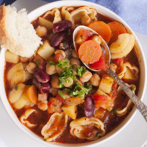 White bowl with a red soup full of red kidney beans, chickpeas, potatoes, carrots and shell pasta. It is topped with fresh parsley. A spoon is taking a carrot.
