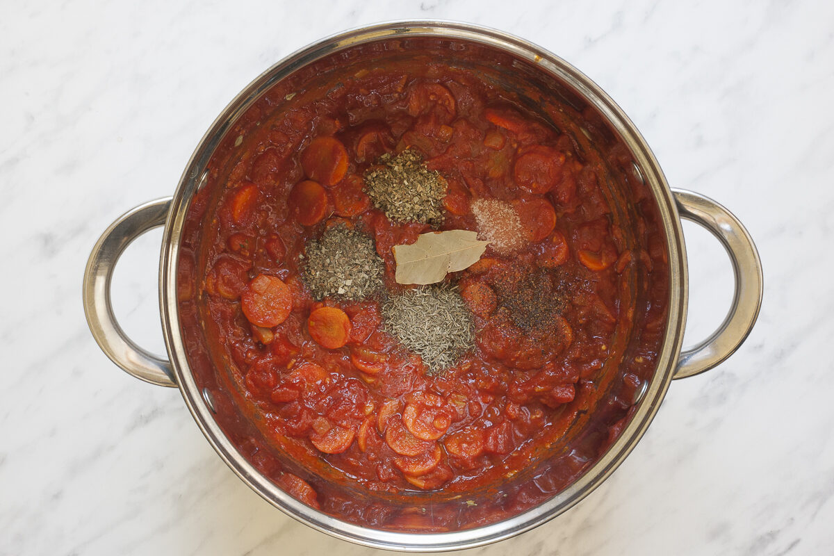 A stockpot with a thick tomato-based soup with sliced vegetables and small heaps of dried herbs.