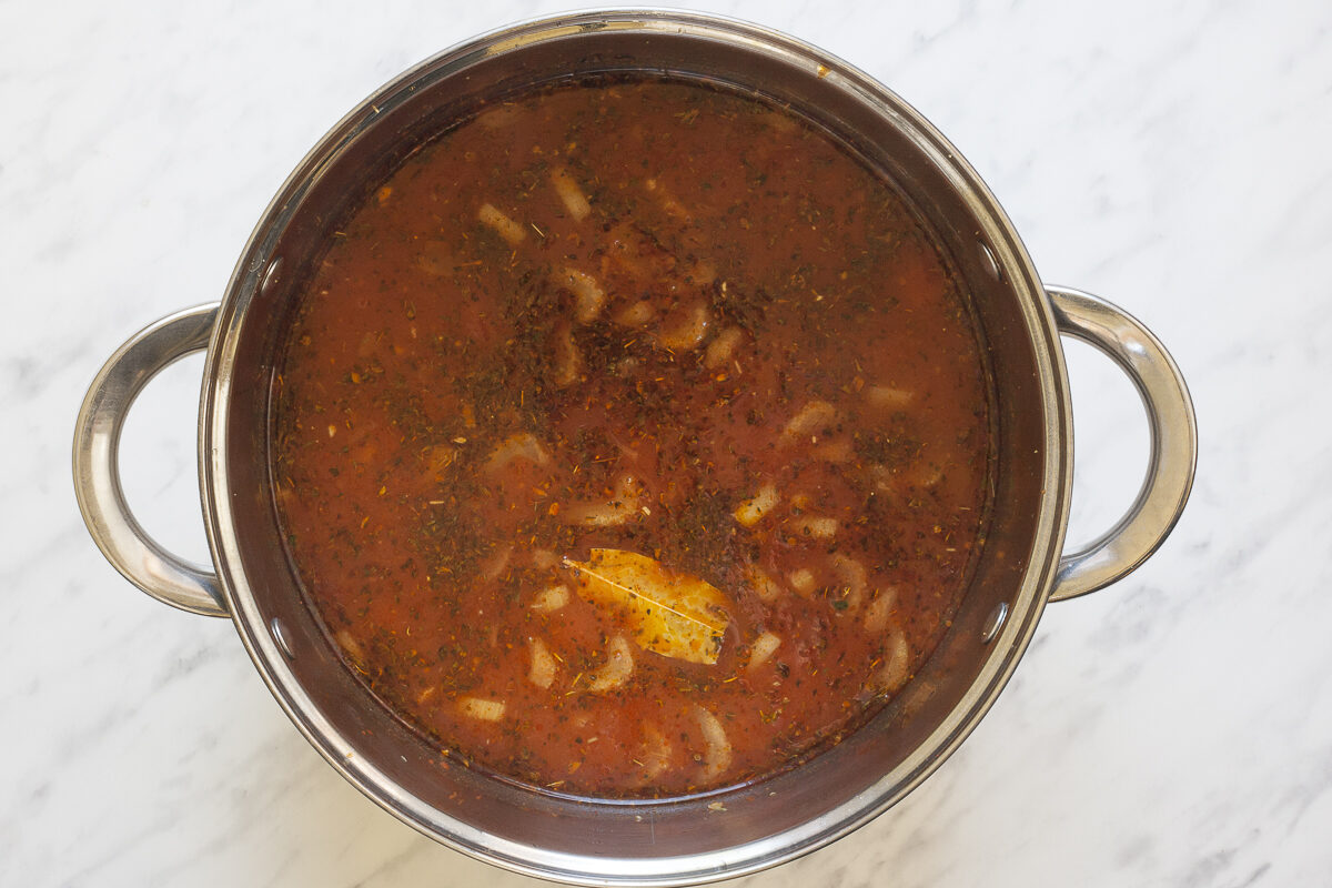 Stockpot with a thin red soup full of sliced vegetables and dried herbs.