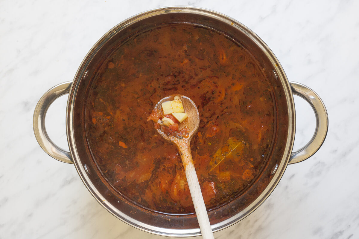 Stockpot with a thin red soup full of sliced vegetables and dried herbs. A wooden spatula shows that diced potatoes were added.