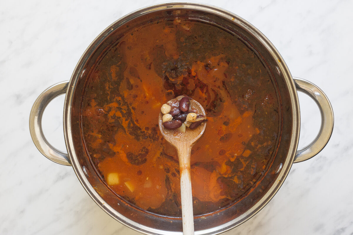 Stockpot with a thin red soup full of sliced vegetables and dried herbs. A wooden spatula shows that beans and chickpeas were added.