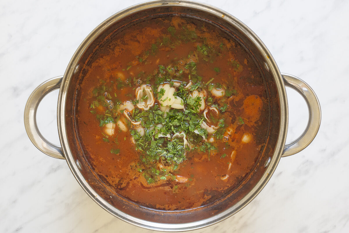 Stockpot with a thin red soup full of sliced vegetables and dried herbs. In the middle shell pasta is visible under a small heap of freshly chopped herbs.