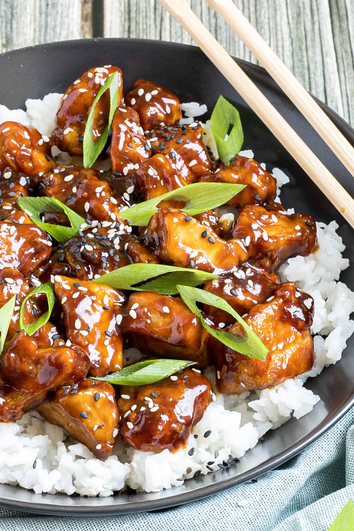 Brown sticky tofu pieces served on top of white rice on a black plate. It is sprinkled with black and white sesame seeds and scallion.