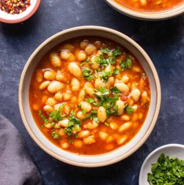 White plate with a red stew and white beans topped with freshly chopped herbs.