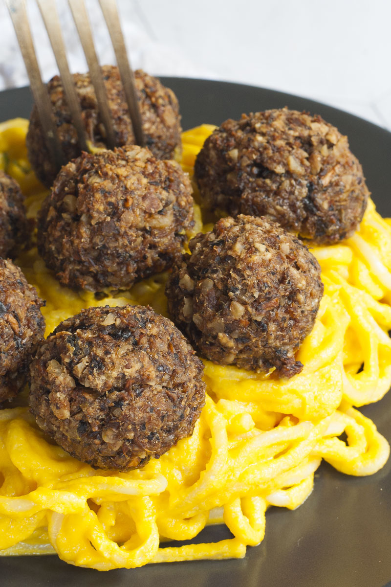 Several mushroom meatballs on top of spaghetti with yellow creamy sauce served on a black plate. A fork is taking on meatballs from the middle.