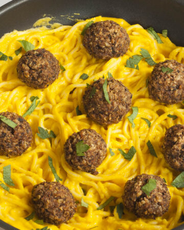 Several mushroom meatballs on top of spaghetti with yellow creamy sauce served on a black plate. A fork is taking on meatballs from the middle.