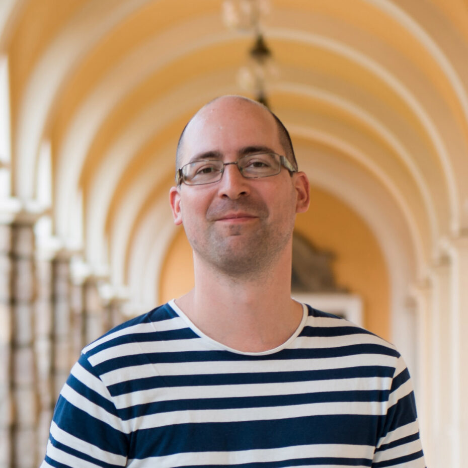 A man with glasses and blue white striped T-shirt in front of yellow archway