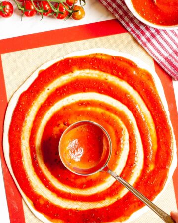 Red pizza sauce is spread in circles on top of a pizza crust with a ladle.