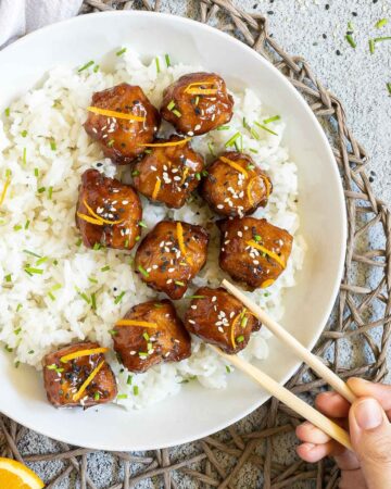 Sticky brown breaded tofu cubes are served on top of rice in a white bowl. It is sprinkled with chopped chives and sesame seeds. A hand is holding chopsticks to take one.