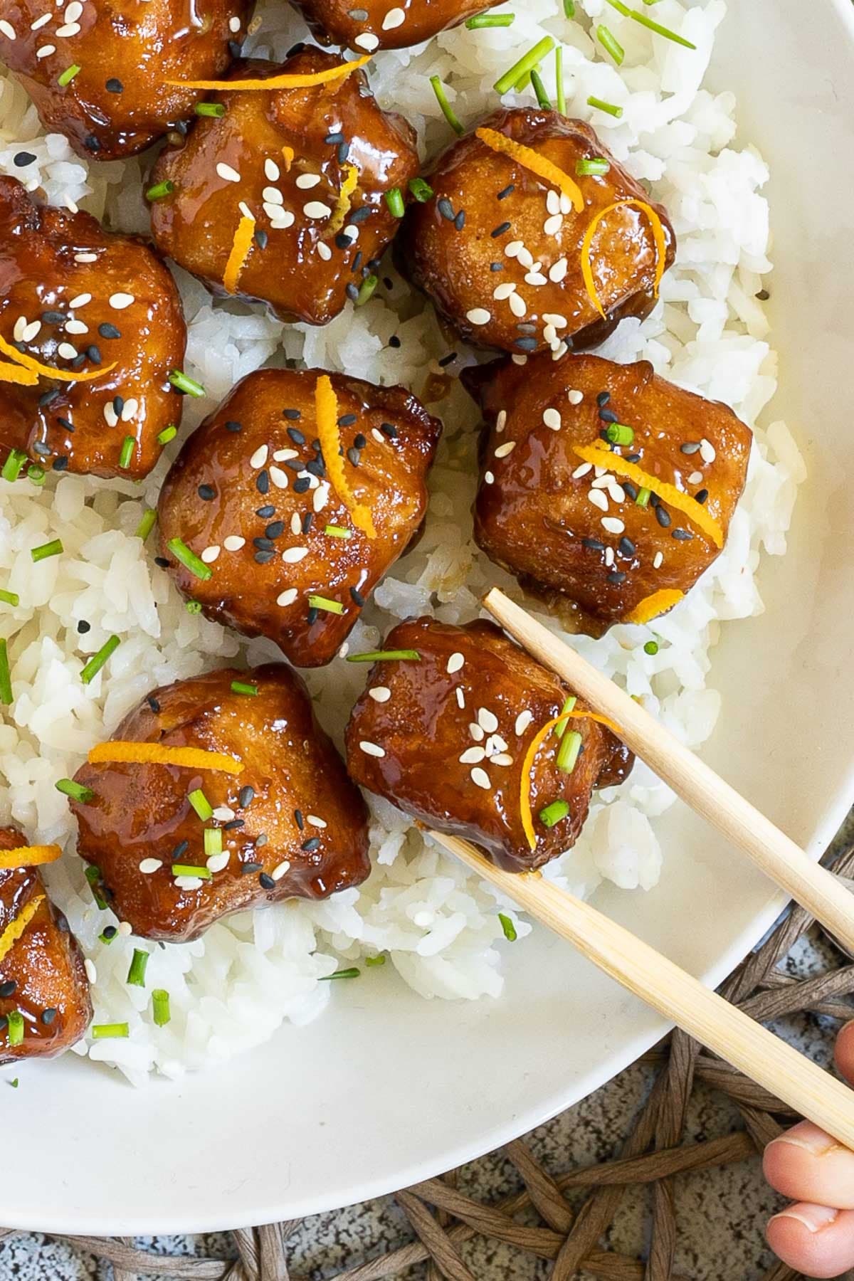 Sticky brown breaded tofu cubes are served on top of rice in a white bowl. It is sprinkled with chopped chives and sesame seeds. A hand is holding chopsticks to take one.