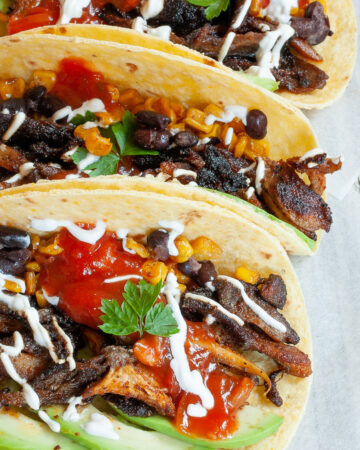 3 small tortillas folded in half on parchment paper filled with mushroom shreds, corn, avocado slices, black beans drizzled with red and white sauce.
