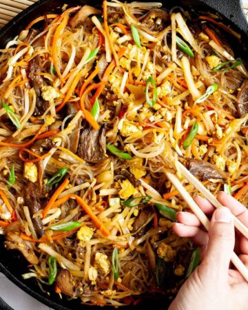 Black skillet from above full of glass noodles, shredded carrots, cabbage, sliced green onion, bean sprouts, mushroom shreds. A hand is holding chopsticks and digging in the middle