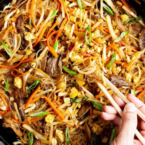Black skillet from above full of glass noodles, shredded carrots, cabbage, sliced green onion, bean sprouts, mushroom shreds. A hand is holding chopsticks and digging in the middle