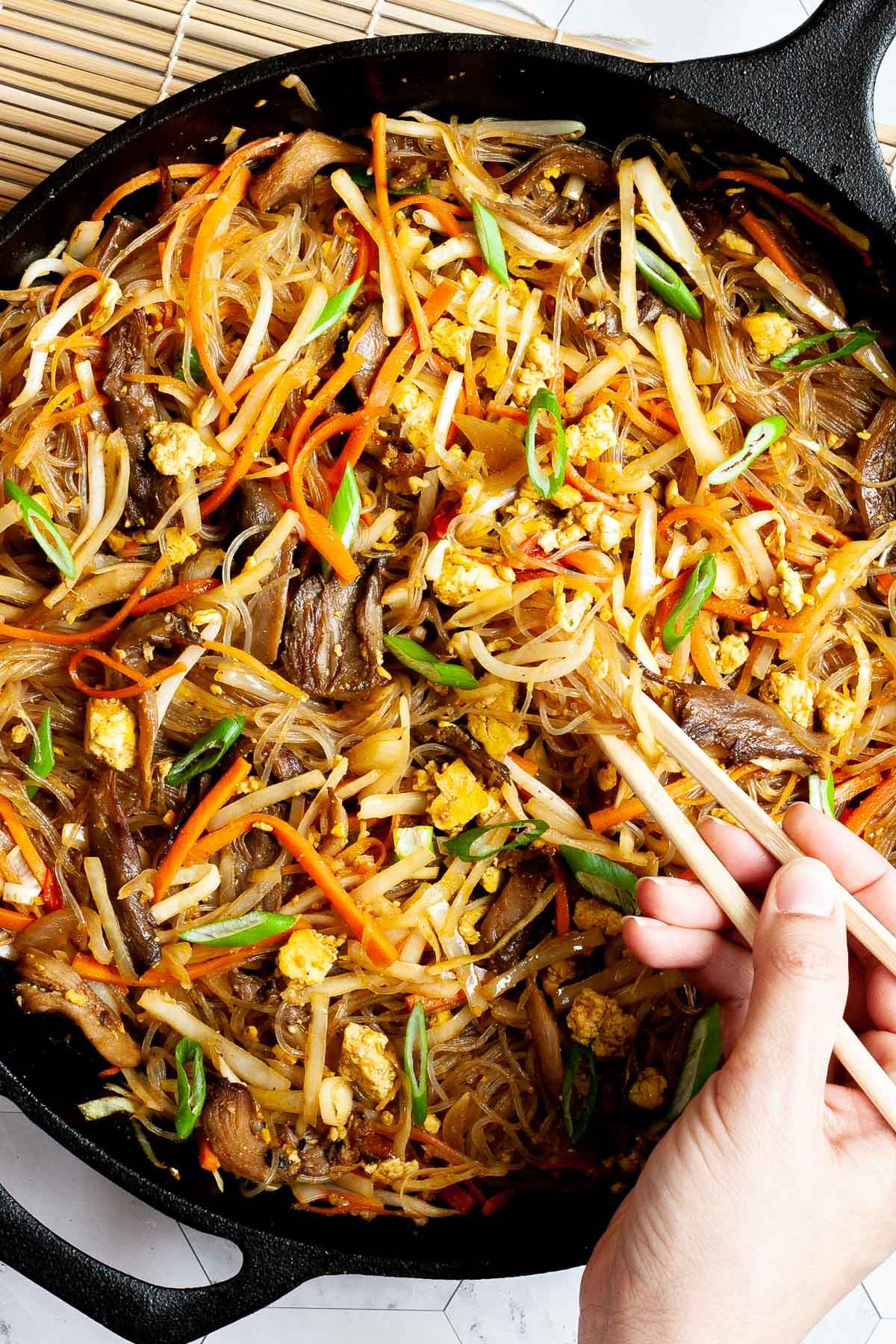 Black skillet from above full of glass noodles, shredded carrots, cabbage, sliced green onion, bean sprouts, mushroom shreds. A hand is holding chopsticks and digging in the middle