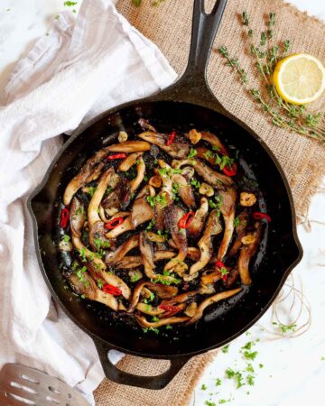 Black cast iron skillet from above with crispy brown oyster mushroom, sliced red pepper, garlic slices and chopped fresh green herbs.