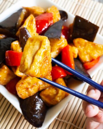 White rectangular plate full of large purple eggplant chunks, puffy brown glazed tofu and red peppers. Blue chopsticks are taking one tofu piece and holding it up in the air.