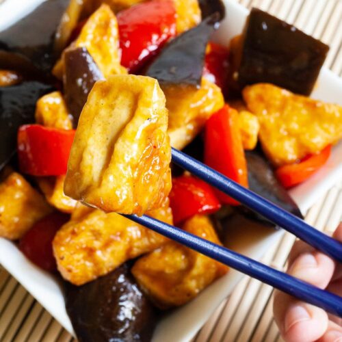 White rectangular plate full of large purple eggplant chunks, puffy brown glazed tofu and red peppers. Blue chopsticks are taking one tofu piece and holding it up in the air.
