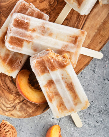 Brown and white popsicles on a wooden cutting board with peach slices scattered around them.