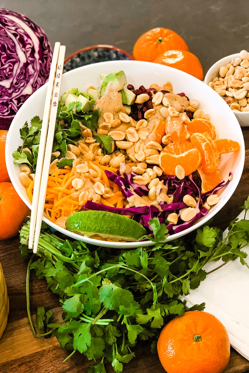 White bowl with peanuts, matchstick carrots, cilantro, purple cabbage, tangerine, and peanut sauce