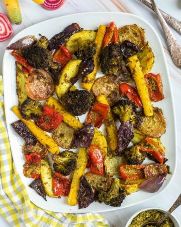 Large white serving plate with vibrant vegetables like broccoli, yellow carrots, red pepper, red onion, pink beet and potato slightly charred around the edges covered in a bit of pesto sauce.