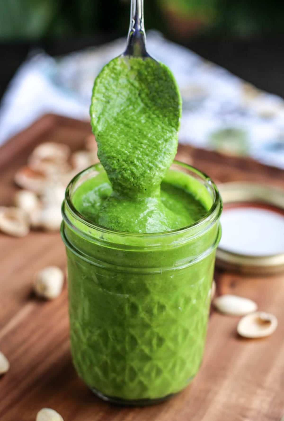 Vibrant green sauce in a glass jar. A spoon is taking from from the top. Pistachios are around it.