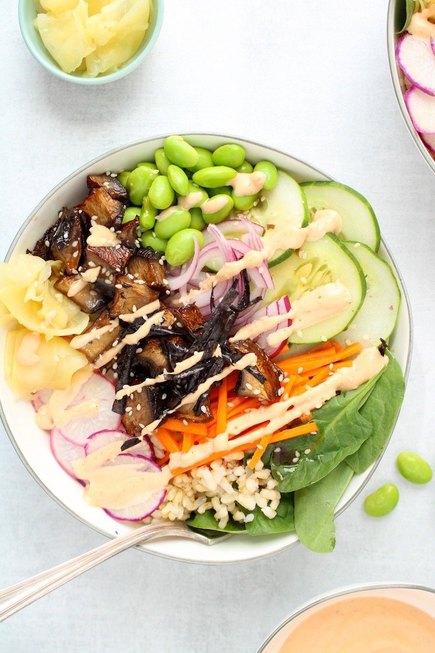 A white bowl from above full of veggies like mushroom slices, spinach leaves, sliced radish, sliced cucumber, matchstick carrots, pruple onion slices and edamame beans drizzled with a light orange colored sauce