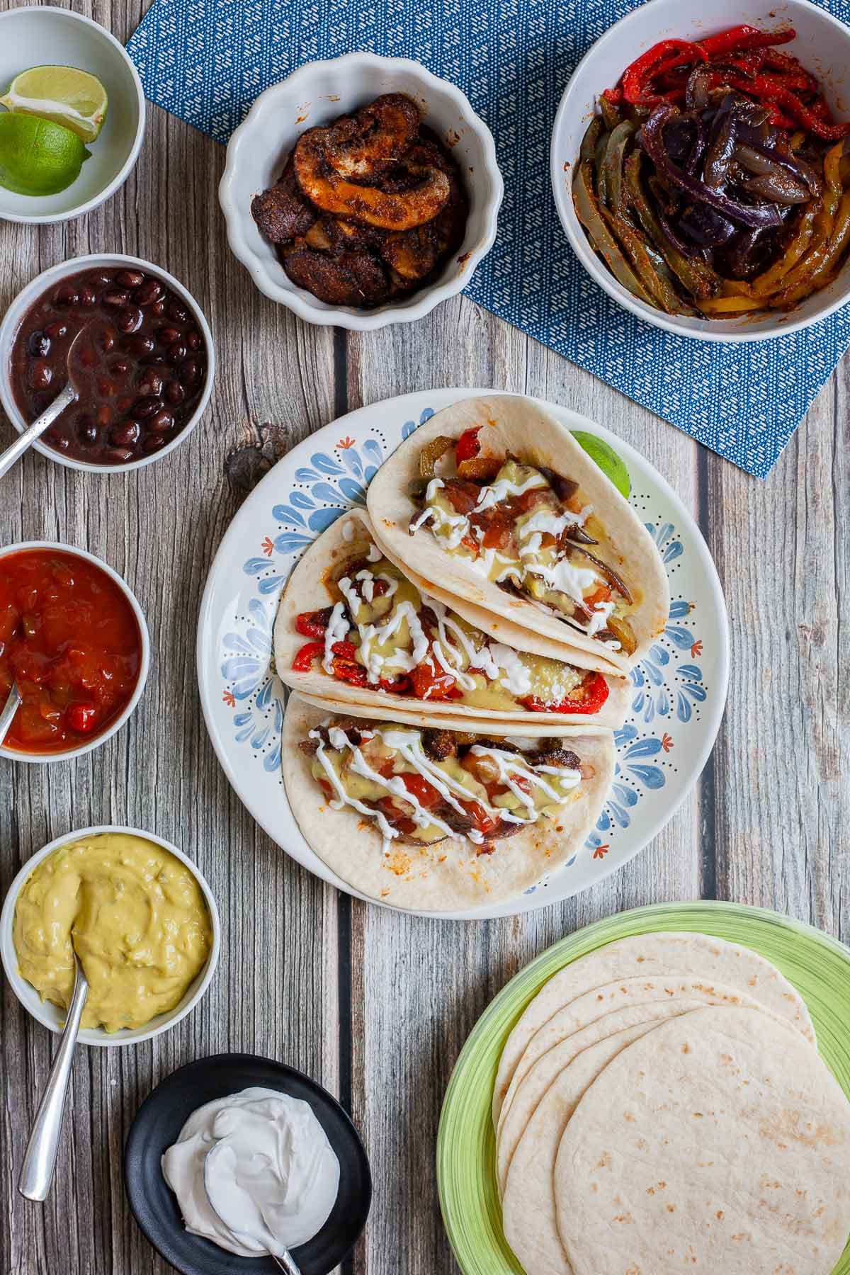 3 soft tortillas folded in half on a white blue plate filled with red pepper slices, purple onion slices and mushroom slices drizzled with white sauce, red salsa and guacamole. Leftover fillings in small bowls are around it. 