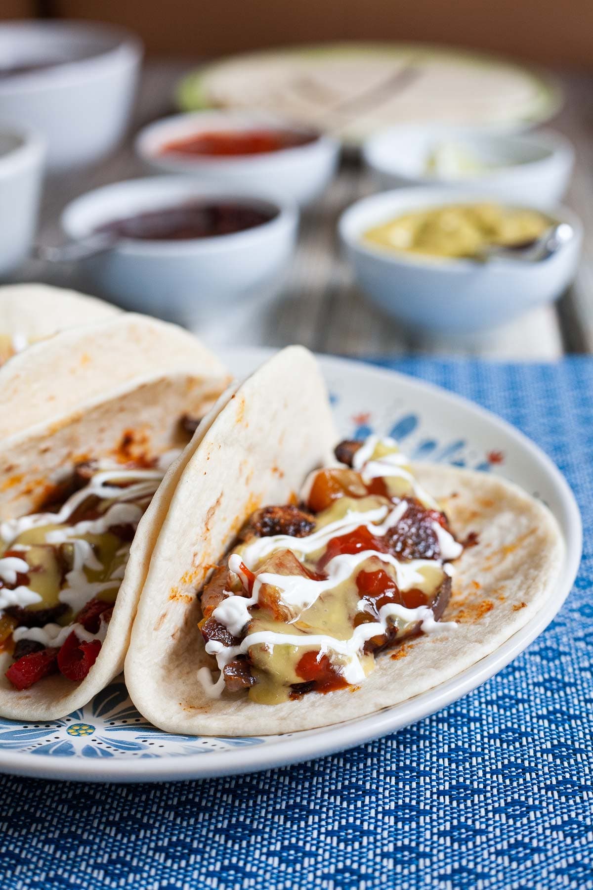 3 soft tortillas folded in half on a white blue plate filled with red pepper slices, purple onion slices and mushroom slices drizzled with white sauce, red salsa and guacamole. Leftover fillings in small bowls are behind it.