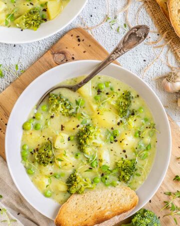 2 white plates of thick yellow stew with potatoes, broccoli, green peas and green herbs. A spoon is placed inside with a toasted slice of bread.