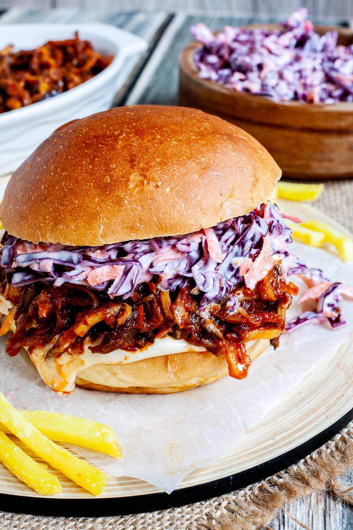 A burger sandwich on a wooden board with white sauce, brown juicy strips and purple cabbage coleslaw. A white bowl is the remaining brown juicy mushroom slices and a wooden bowl with more coleslaw salad and french fries