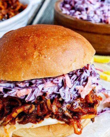 A burger sandwich on a wooden board with white sauce, brown juicy strips and purple cabbage coleslaw. A white bowl is the remaining brown juicy mushroom slices and a wooden bowl with more coleslaw salad and french fries