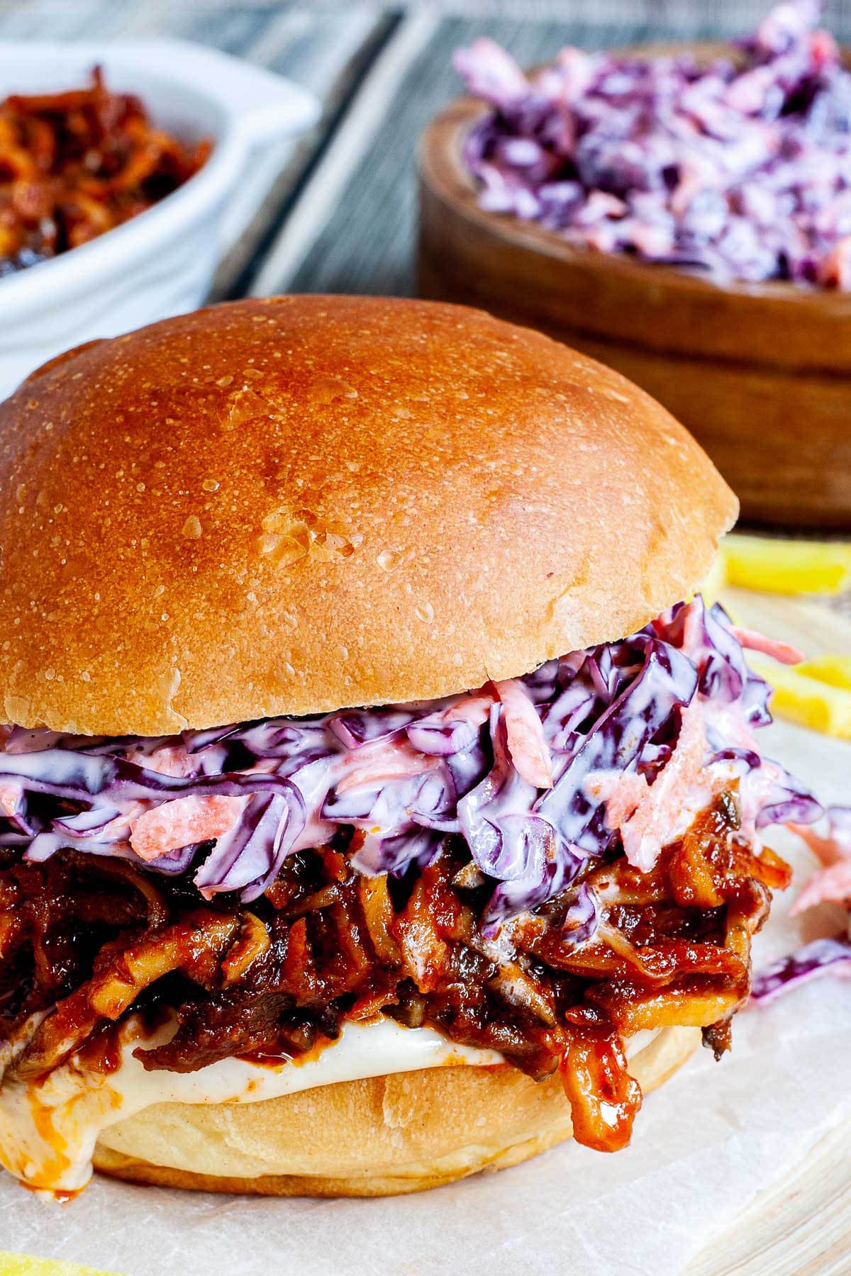 A burger sandwich on a wooden board with white sauce, brown juicy strips and purple cabbage coleslaw. A white bowl is the remaining brown juicy mushroom slices and a wooden bowl with more coleslaw salad and french fries