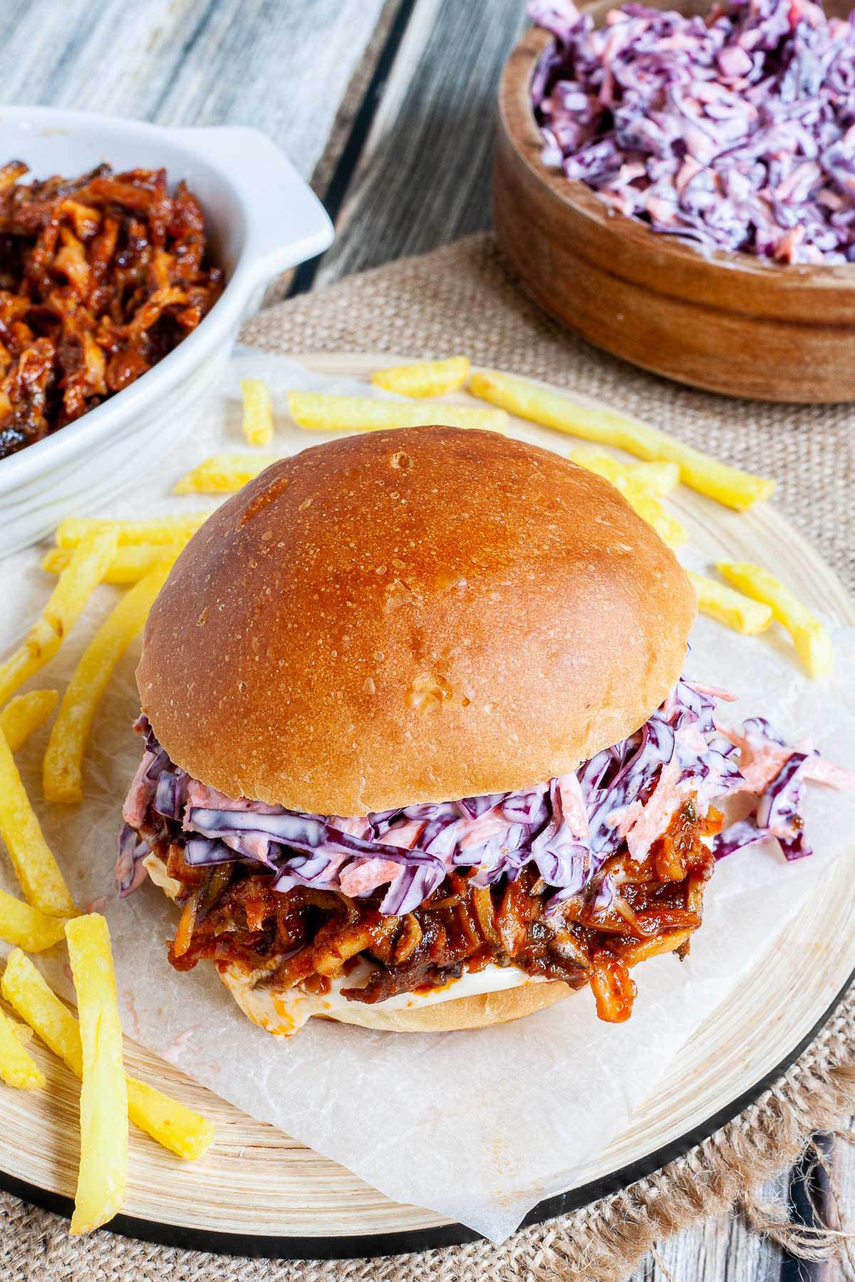 A burger sandwich on a wooden board with white sauce, brown juicy strips and purple cabbage coleslaw. A white bowl is the remaining brown juicy mushroom slices and a wooden bowl with more coleslaw salad and french fries