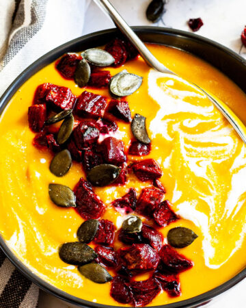 Yellow cream soup in a black bowl topped with diced roasted beetroot, green pumpkin seeds and drizzled with a white sauce.