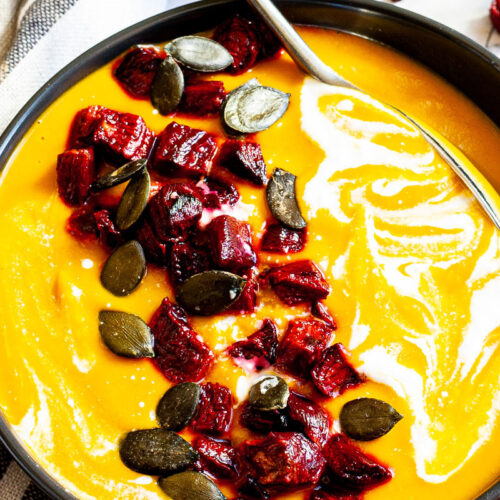 Yellow cream soup in a black bowl topped with diced roasted beetroot, green pumpkin seeds and drizzled with a white sauce.