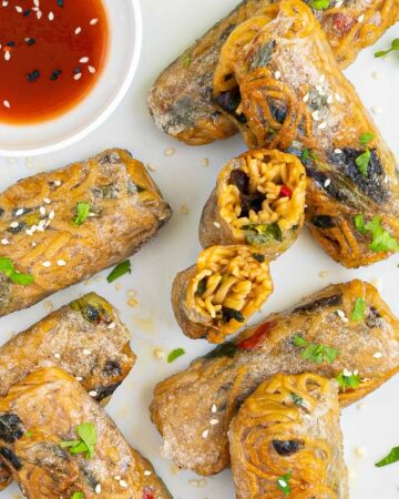 Several ramen spring rolls are on a white plate. A red sweet chili sauce is placed in a small white bowl right next to them. They are also sprinkled with chopped green herbs. 2 rolls are cut in half so the yellow noodles and some green and brown pieces are visible.