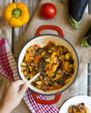 A red pot from above with veggie chunks of all colors: red, yellow, green, orange, brown. There is a serving spoon taking a portion from the middle.
