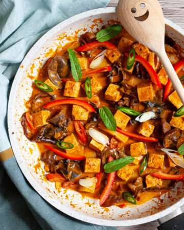 White frying pan with a darkbrown red sauce tofu cubes, sliced bell peppers, sliced green chilies, mint leaves, sliced spring onion, mushrooms slices.