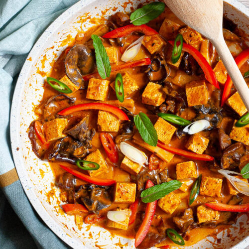 White frying pan with a darkbrown red sauce tofu cubes, sliced bell peppers, sliced green chilies, mint leaves, sliced spring onion, mushrooms slices.