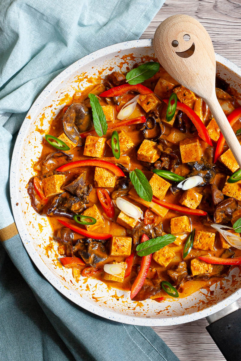 White frying pan with a darkbrown red sauce tofu cubes, sliced bell peppers, sliced green chilies, mint leaves, sliced spring onion, mushrooms slices.