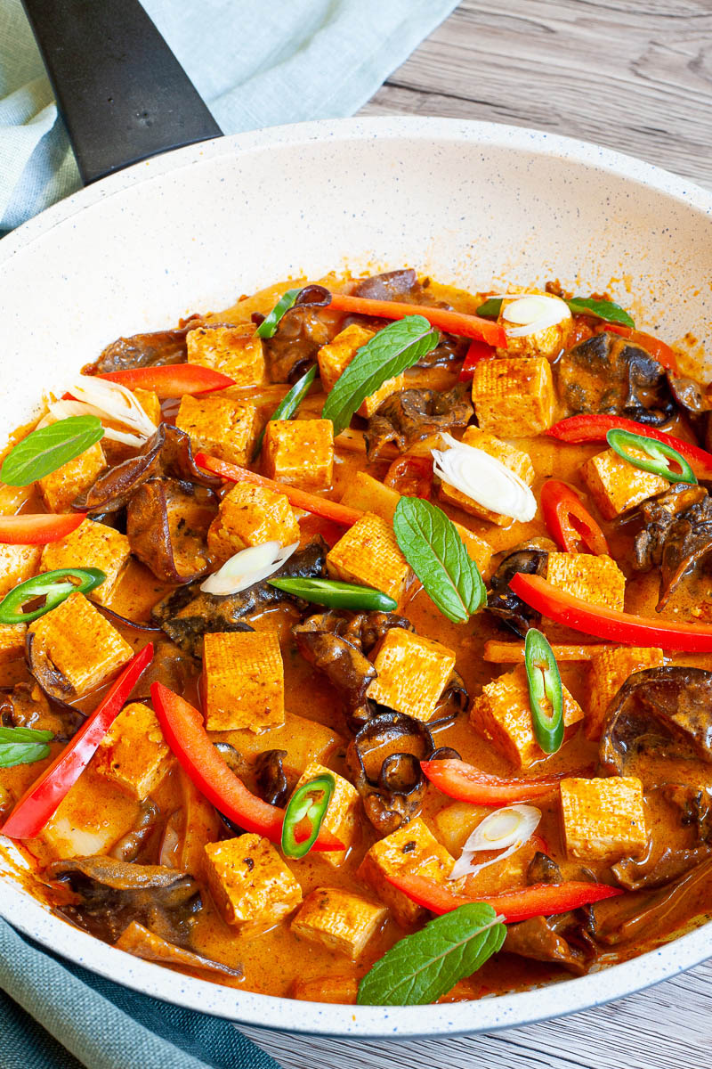 White frying pan with a darkbrown red sauce tofu cubes, sliced bell peppers, sliced green chilies, mint leaves, sliced spring onion, mushrooms slices.