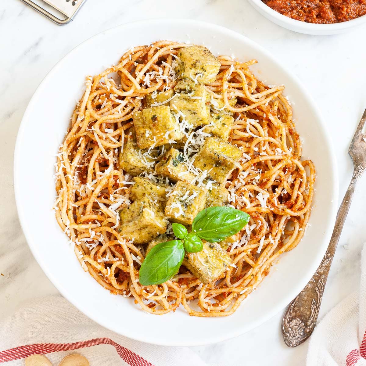 Large white plate with red pesto spaghetti topped with green-yellow tofu cubes, shredded cheese and basil leaves.