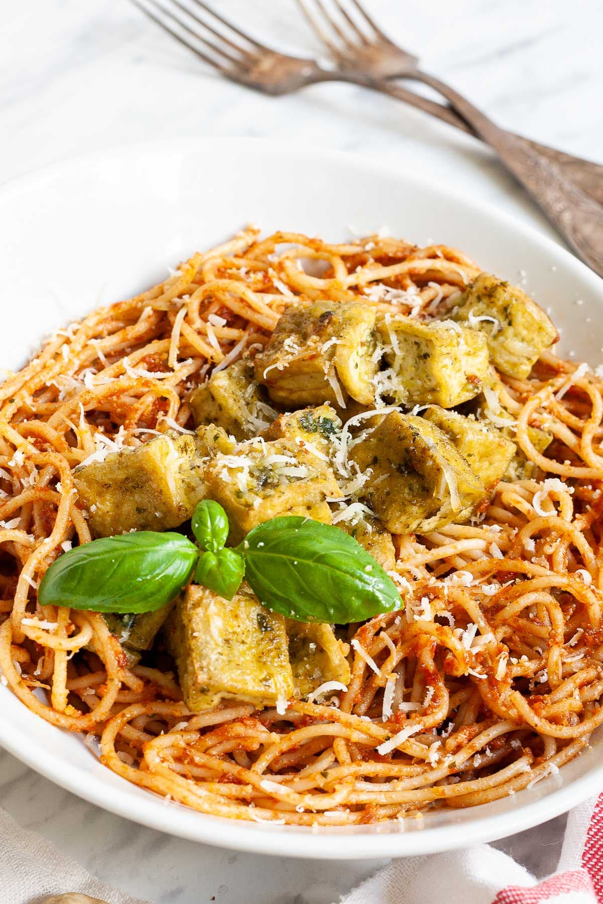 Large white plate with red pesto spaghetti topped with green-yellow tofu cubes, shredded cheese and basil leaves.
