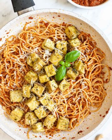 Large white frying pan with red pesto spaghetti topped with green-yellow tofu cubes, shredded cheese and basil leaves.