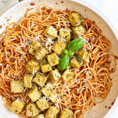 Large white frying pan with red pesto spaghetti topped with green-yellow tofu cubes, shredded cheese and basil leaves.