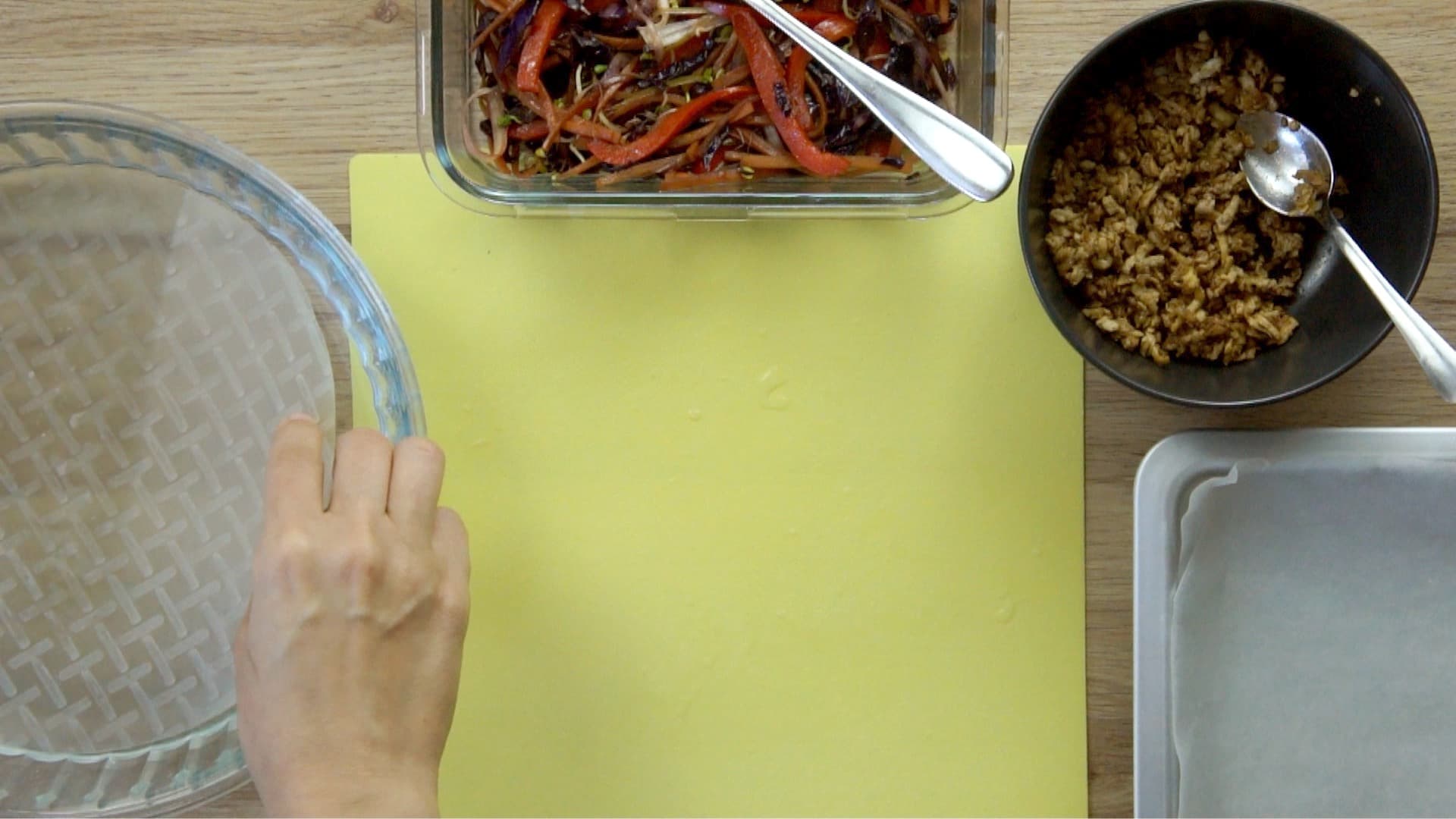 A hand submerges a rice paper in shallow water. The fillings are in small bowls next to a yellow board.