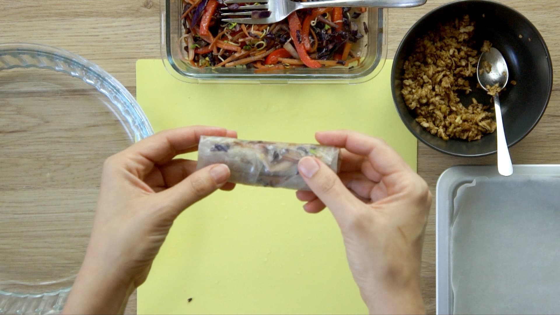 A hand is holding a rolled rice paper . A hand is folding the left side of the rice paper over half of the filling. The fillings are in small bowls next to it.